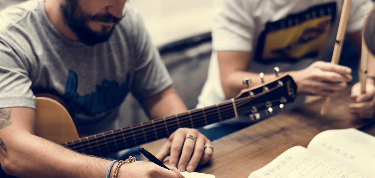 two men at the table. one of them is writing and holding a guitar on his lap.